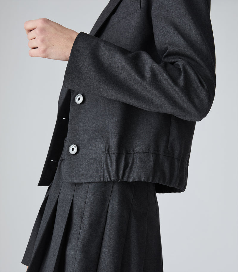Close-up of a person wearing a dark charcoal gray tailored blazer with silver buttons and a matching pleated skirt against a light gray studio background.