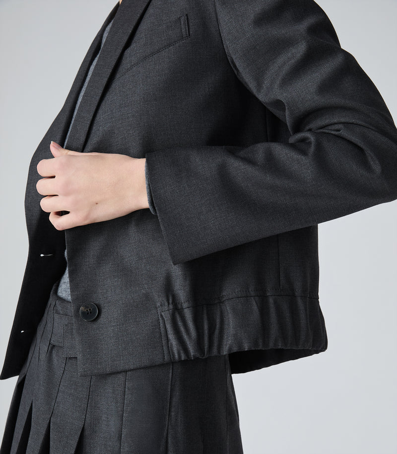 Close-up of a person wearing a dark charcoal gray tailored blazer with structured shoulders and a single visible black button, paired with matching dark pleated trousers against a light gray studio background.