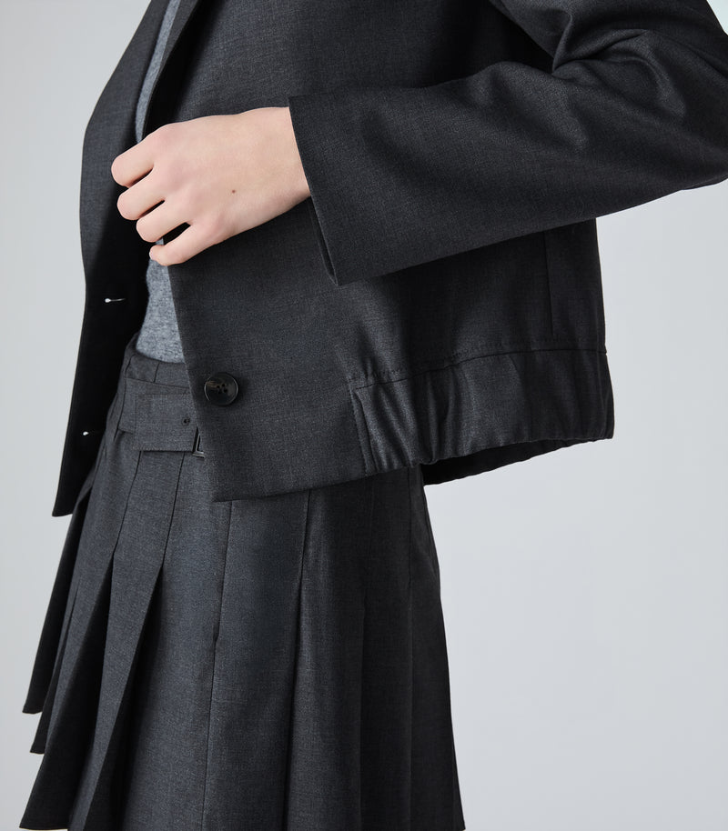 A close-up of a model wearing a dark charcoal gray tailored jacket with a single visible button and pleated skirt, against a light gray studio background.