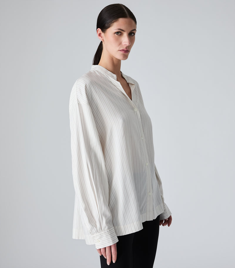 Studio product view of a model wearing an oversized, ivory-beige pinstripe boyfriend shirt. The image showcases the relaxed drape, dropped shoulder seams, a banded collar buttoned halfway to create a subtle V-neckline, and long sleeves with structured cuffs, paired with black trousers against a seamless light grey background.