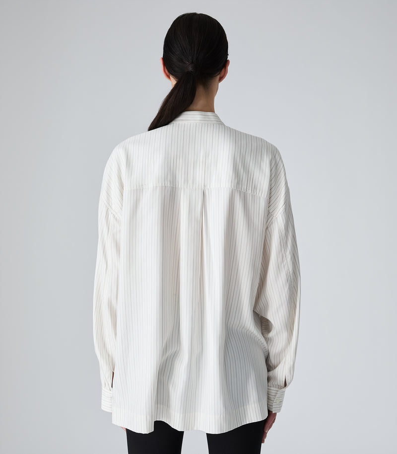 Full-length rear studio view of a model showcasing an oversized ivory-beige pinstripe boyfriend shirt. The image highlights a structured back yoke with a prominent centre-back box pleat that enhances the shirt's fluid drape, dropped shoulder seams, a clean band collar, and long sleeves with buttoned cuffs, set against a seamless light grey backdrop.