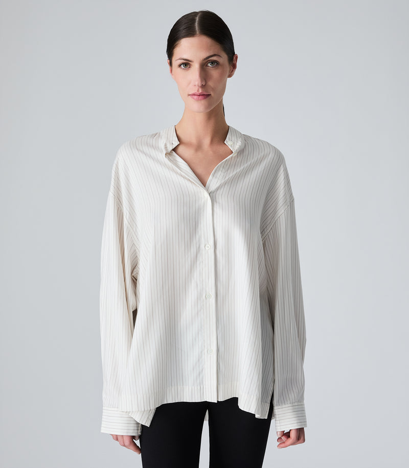 Studio product view from a three-quarter angle, focusing on a model wearing an oversized, ivory-beige pinstripe boyfriend shirt. The image highlights the relaxed drape, dropped shoulders, a casual band collar styled open to create a V-neckline, and long volume sleeves with structured cuffs, paired with black trousers against a seamless light grey background.