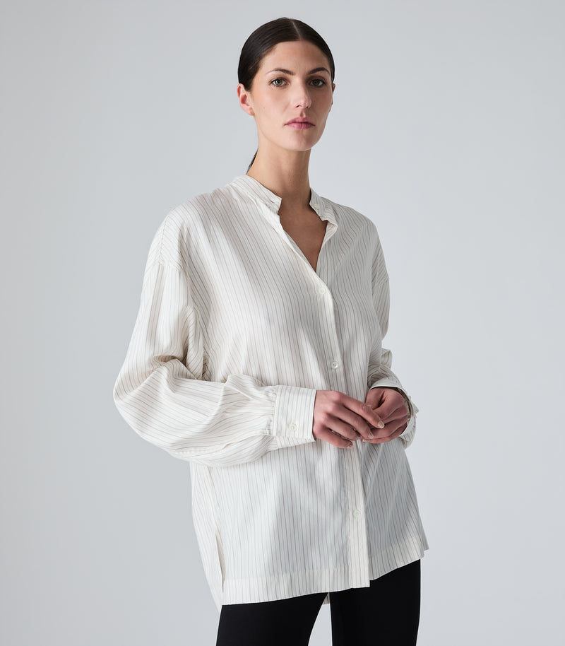 Studio product view from a three-quarter angle, focusing on a model wearing an oversized, ivory-beige pinstripe boyfriend shirt. The image highlights the relaxed drape, dropped shoulders, a casual band collar styled open to create a V-neckline, and long volume sleeves with structured cuffs, paired with black trousers against a seamless light grey background.