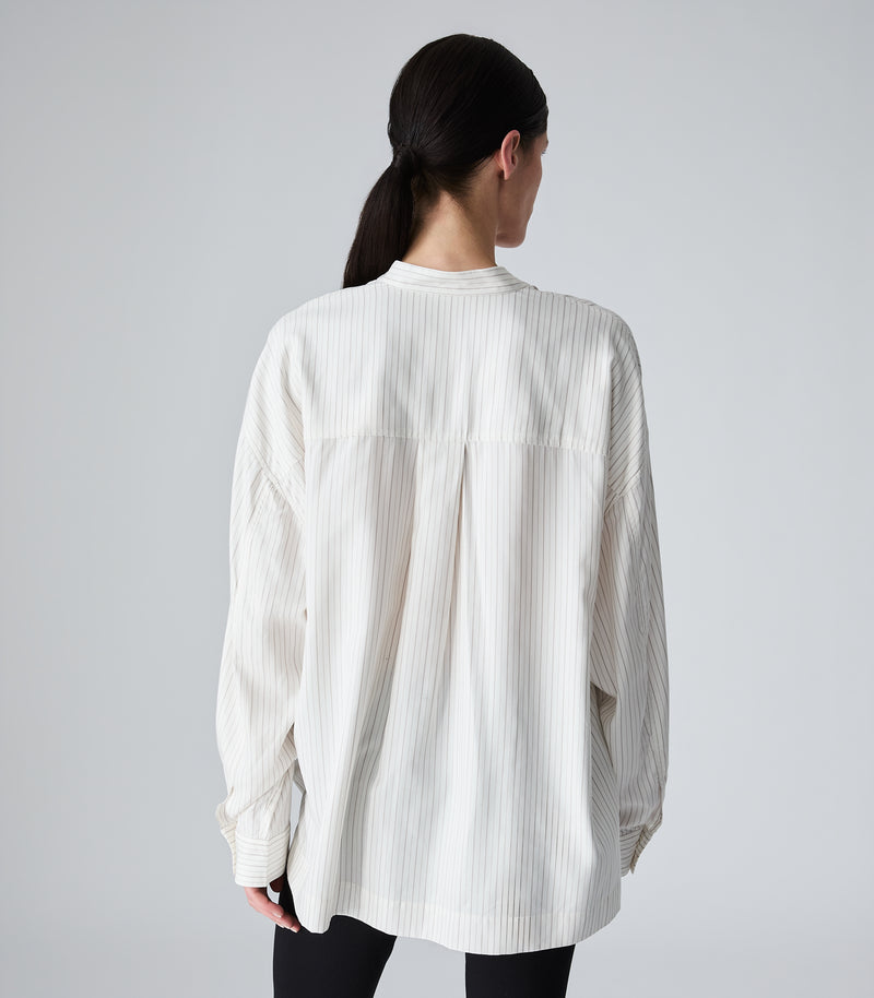Three-quarter length rear studio view of a model showcasing an oversized ivory-beige pinstripe boyfriend shirt. The image highlights a structured back yoke with a prominent centre-back box pleat that enhances the shirt's fluid drape, dropped shoulder seams, a clean band collar, and long sleeves with buttoned cuffs, set against a seamless light grey backdrop.