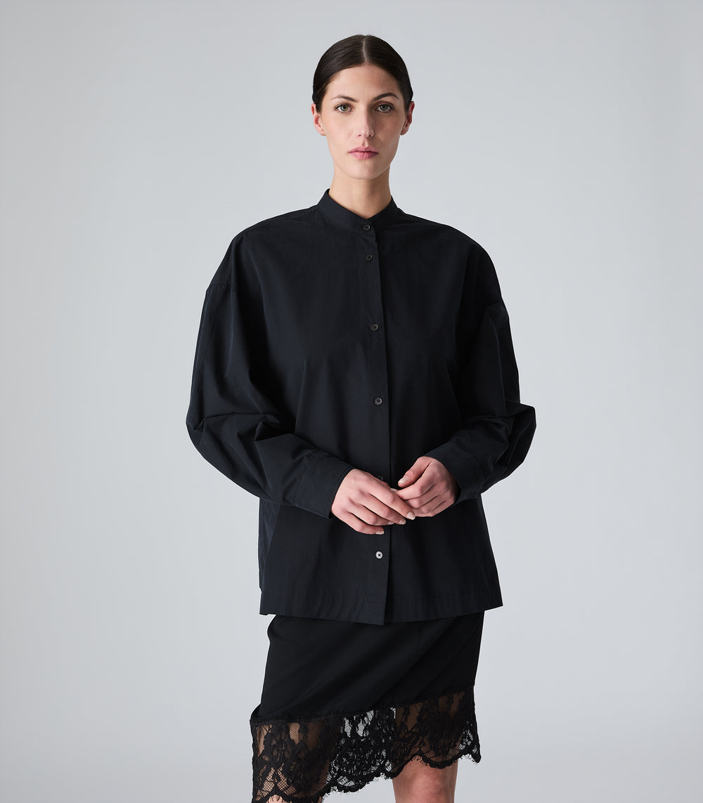 A model wearing a black long-sleeve button-up shirt with a band collar and a black skirt featuring a lace hem, photographed against a neutral light gray background.