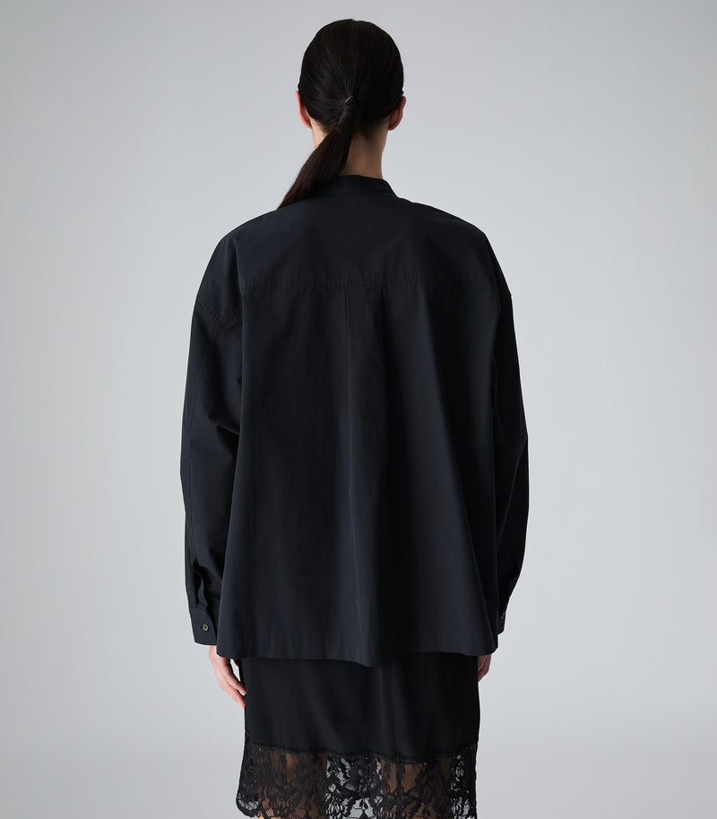 A model shown from the back wearing an oversized black long-sleeve shirt or jacket over a black lace-trimmed garment, photographed against a plain light gray studio background.