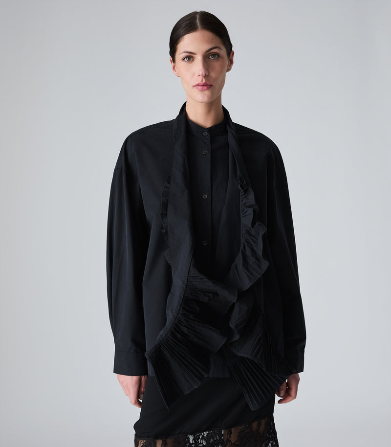 A model stands against a light gray studio background wearing a black long-sleeve draped shirt jacket with a high collar, a black scarf with dramatic pleated ruffle trim on both ends, paired with a black lace-trimmed mini skirt.