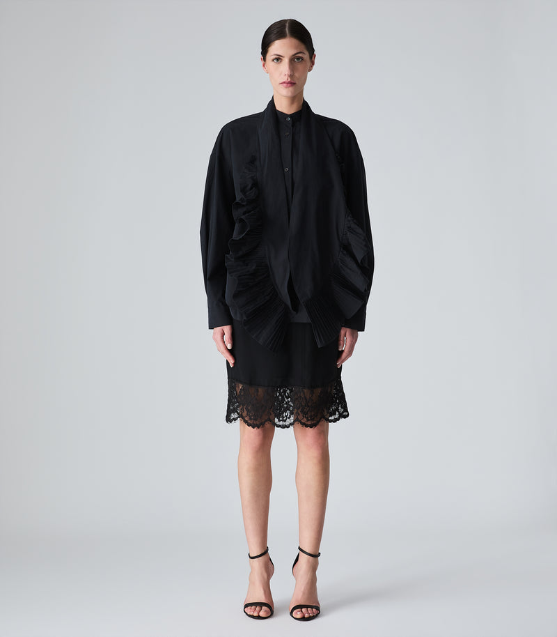 A model stands against a light gray studio background wearing a black long-sleeve draped shirt jacket with a high collar, a black scarf with dramatic pleated ruffle trim on both ends, paired with a black lace-trimmed mini skirt and black strappy heels.