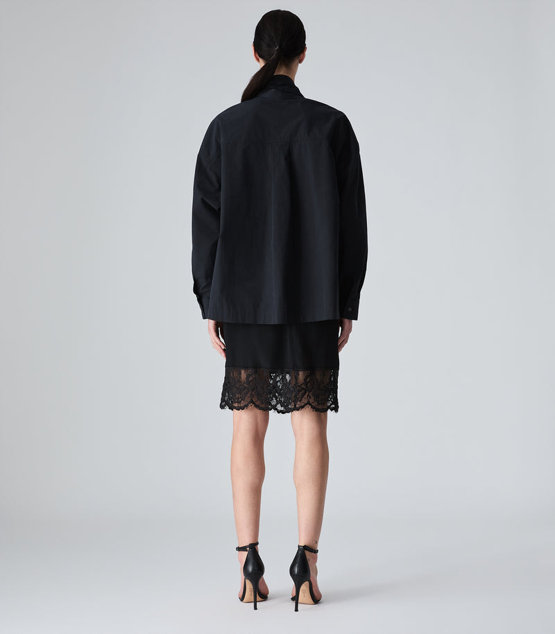 A model shown from the back wearing a black oversized long-sleeve shirt and a black mini skirt with a lace hem, paired with black high-heeled sandals, against a light gray studio background.