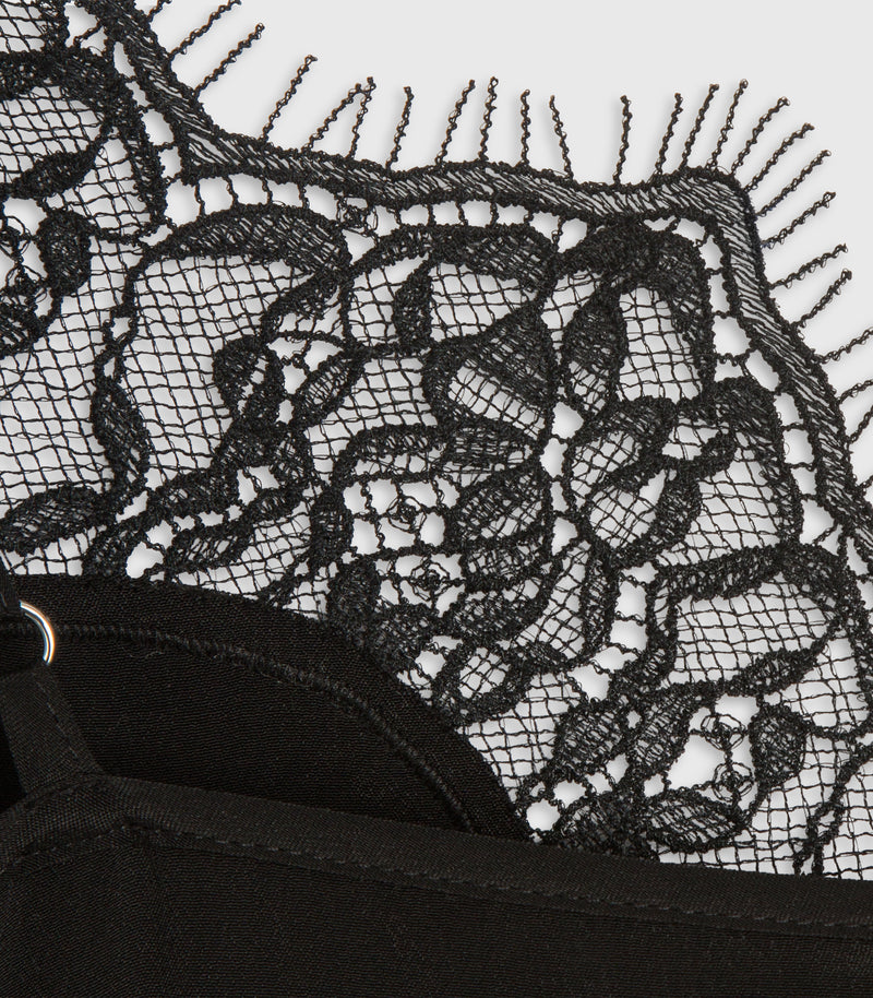 Close-up of a black lace garment with floral lace pattern and fringe trim over a black fabric base, shown on a plain light background.