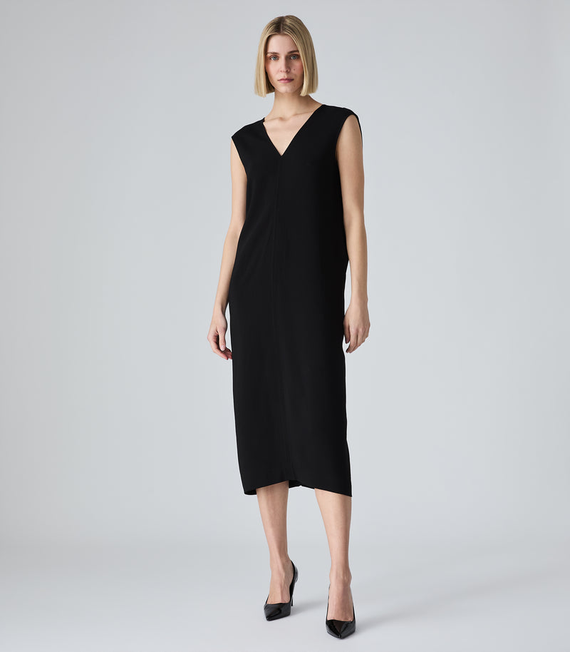 A woman modeling a sleeveless black midi dress with a V-neckline, paired with black pointed-toe heels, against a light gray studio background.