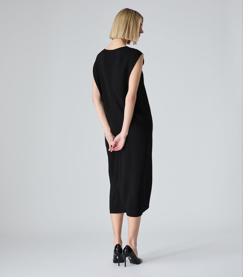 A woman with short blonde hair is shown from the back wearing a sleeveless black midi dress and black heels, standing against a light gray studio background.