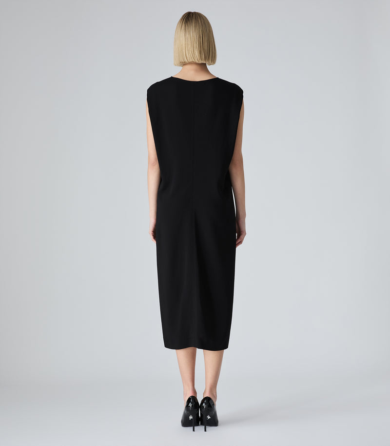 A woman with a short blonde bob is shown from the back wearing a sleeveless black midi dress and black pointed-toe heels against a light gray studio background.
