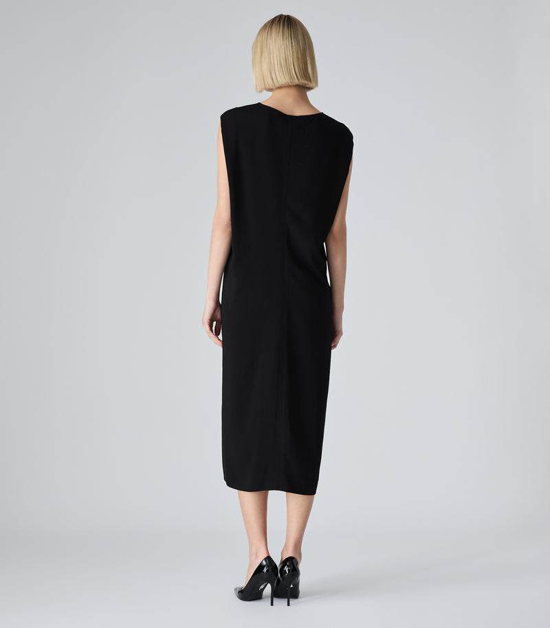 A woman with short blonde hair is shown from the back wearing a sleeveless black midi dress and black heels against a light gray studio background.