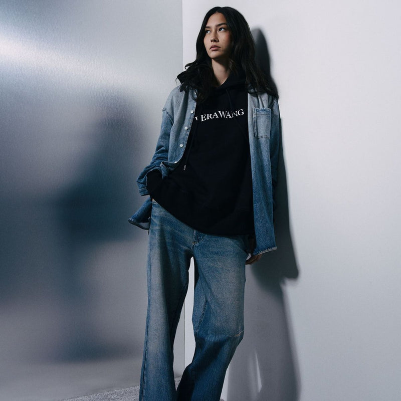 A model leans against a white wall wearing a black graphic hoodie with 'BRAWANG' printed in white across the chest, layered under a light blue denim jacket and paired with wide-leg blue jeans.