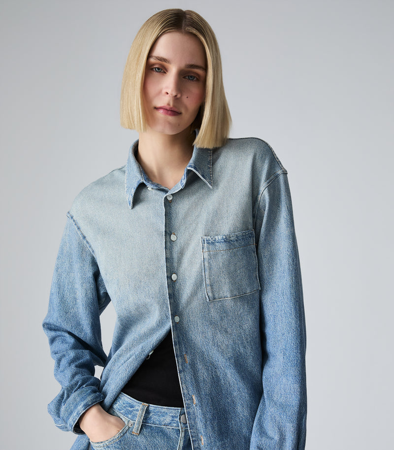 A woman with a short blonde bob wearing a light-wash blue denim shirt-jacket over a black top, photographed against a plain light gray studio background.