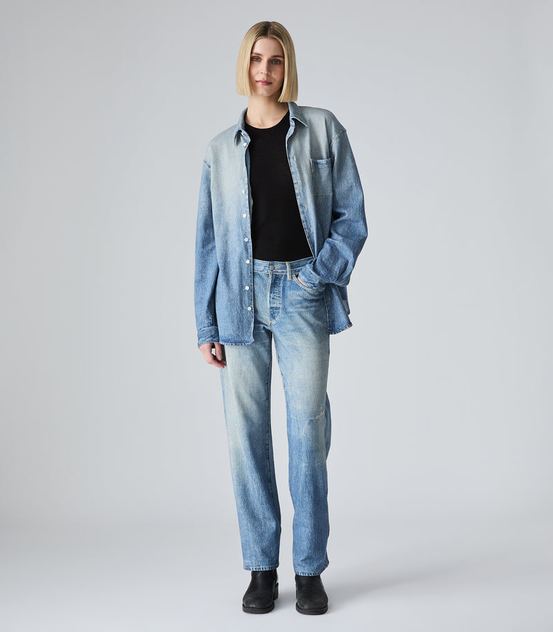 A model stands against a light gray studio background wearing a light-wash blue denim button-up shirt, matching light-wash blue jeans, a black crew-neck top, and black boots.
