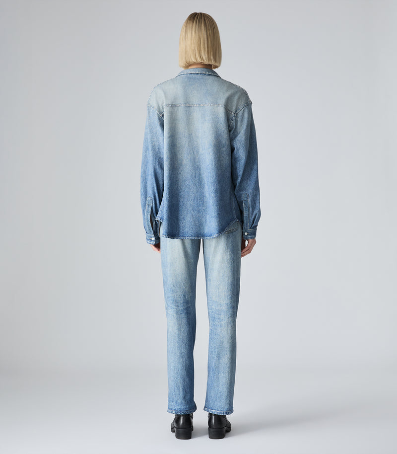 A model viewed from the back wearing a light blue denim shirt-jacket and matching light blue denim jeans, standing against a neutral light gray studio background.