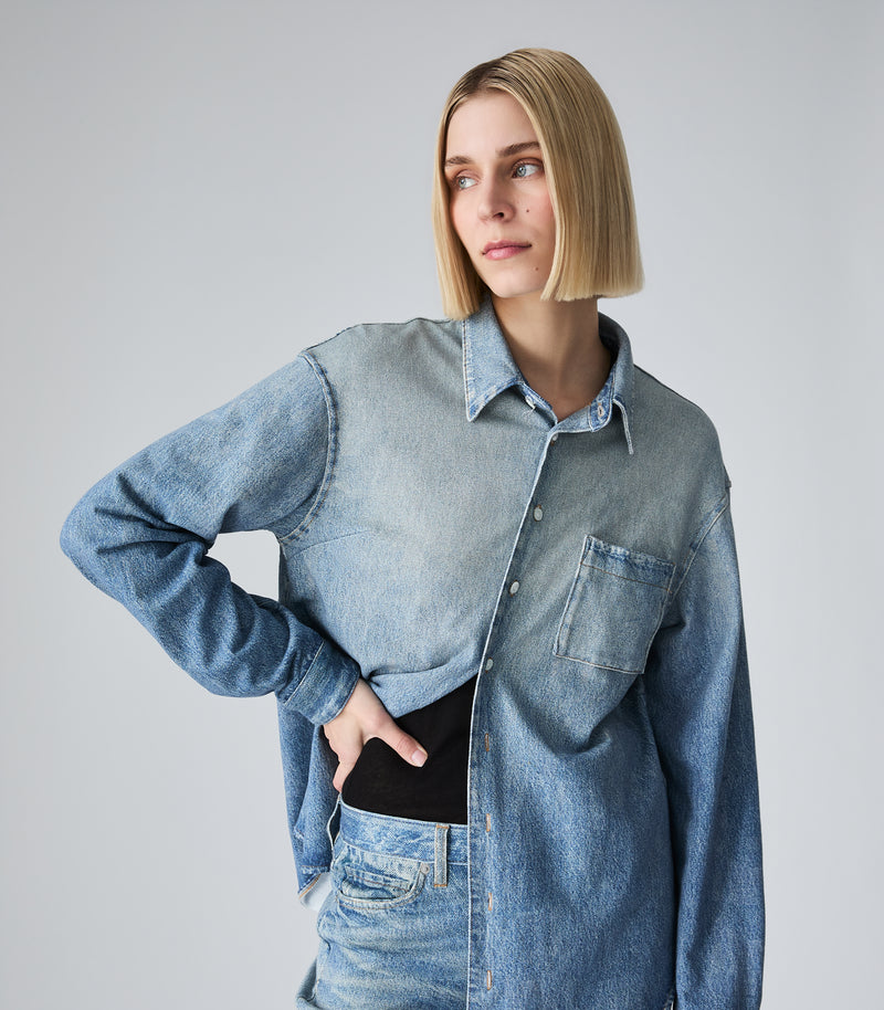 A woman with a short blonde bob wearing a light blue denim shirt-jacket over a black top and matching denim skirt, posing against a neutral gray studio background.