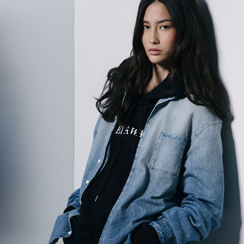 A model with long dark hair is wearing a light blue denim button-up shirt over a black hoodie with white text, leaning against a light gray wall.