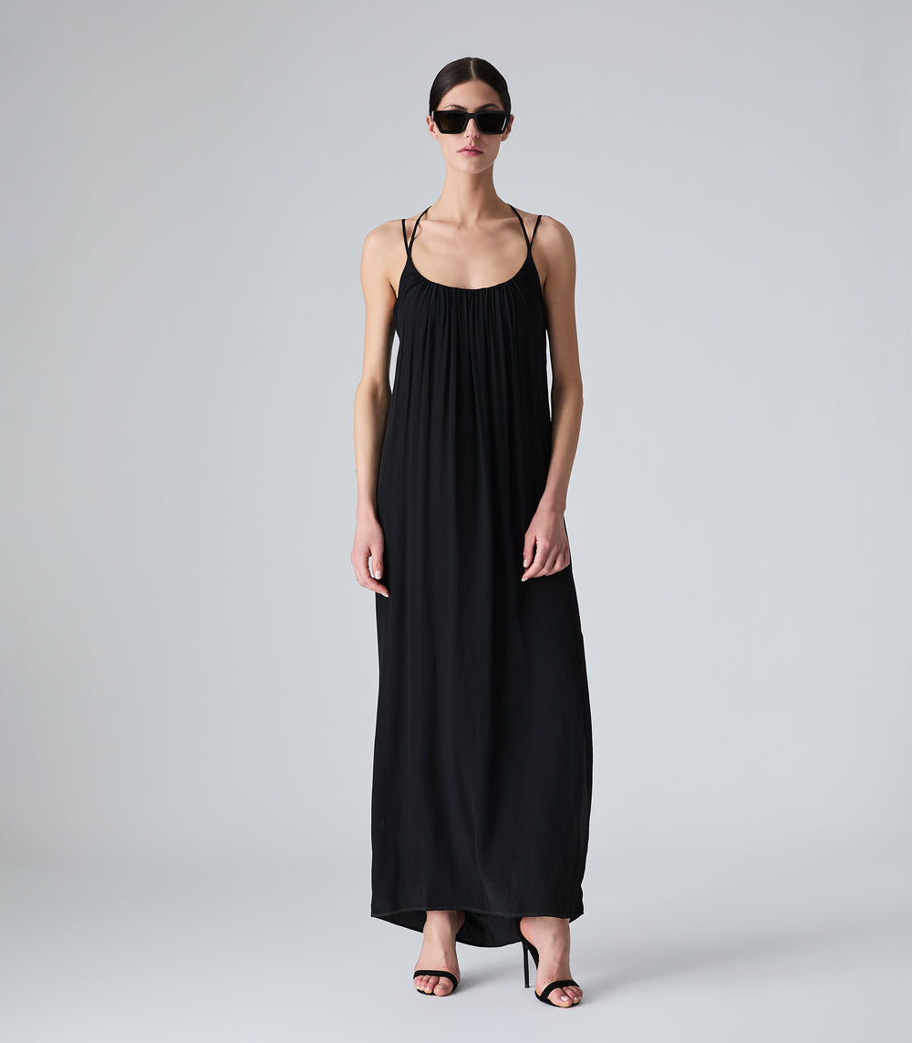 Full-length view of a model wearing a fluid maxi-length black slip dress, featuring a gathered scoop neckline, delicate double spaghetti straps, and a relaxed silhouette. The look is completed with black shield sunglasses and black strappy stiletto sandals against a light grey background.