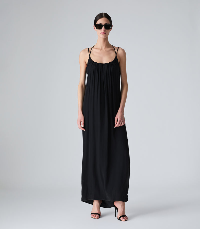 Full-length view of a model wearing a fluid maxi-length black slip dress, featuring a gathered scoop neckline, delicate double spaghetti straps, and a relaxed silhouette. The look is completed with black shield sunglasses and black strappy stiletto sandals against a light grey background.