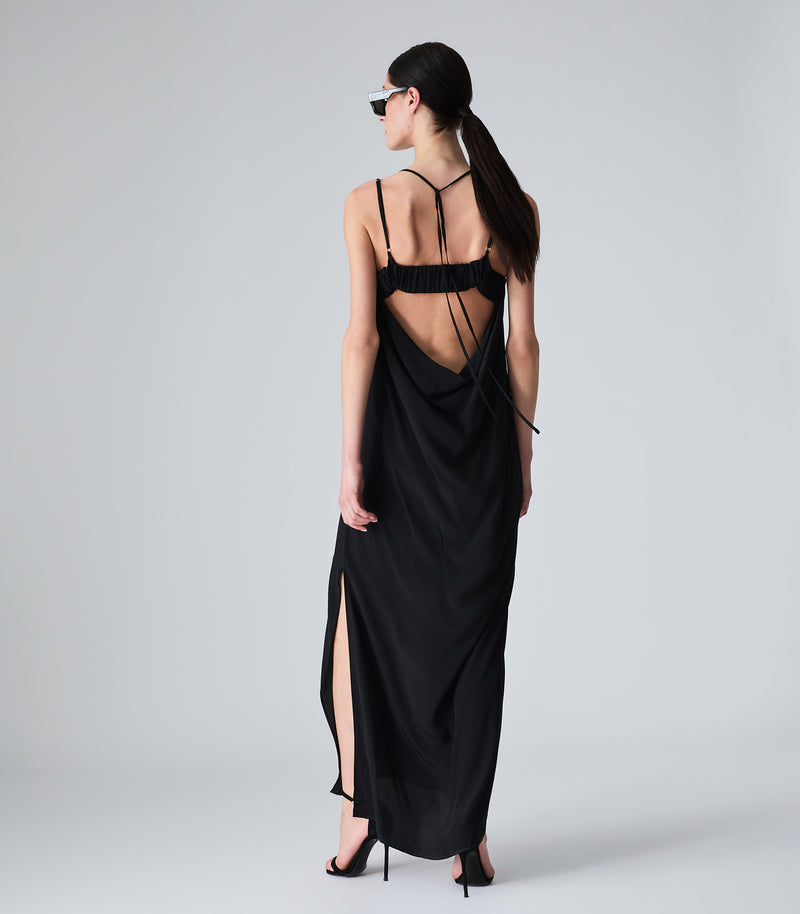 Full-length back view of a model wearing a fluid black maxi slip dress, showcasing a dramatic open back with a gathered upper strap, a low cowl drape, thin ties, and a subtle side leg slit. The look is styled with black stiletto heeled sandals against a light grey background.