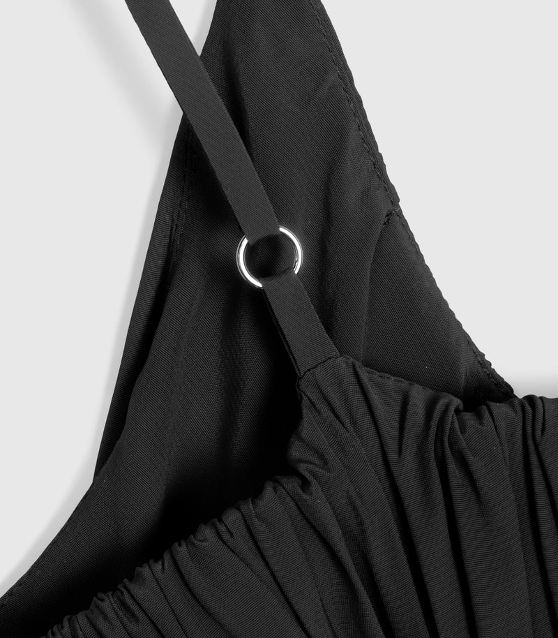 Close-up product view of a black slip dress, showcasing a silver metal O-ring hardware detail connecting the delicate shoulder strap to a finely gathered, puckered bodice. The shot highlights the dense, premium textile weave and precision tonal stitching against a light grey background.