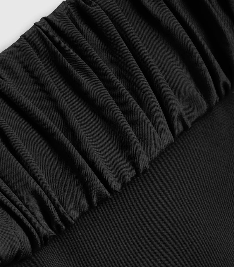 Macro close-up product view of a black slip dress, showcasing a thick, tightly gathered ruched elastic strap stretching diagonally across smooth, lightweight matte jersey fabric. The shot highlights the dense, uniform texture of the folds and premium textile construction against a light grey background.