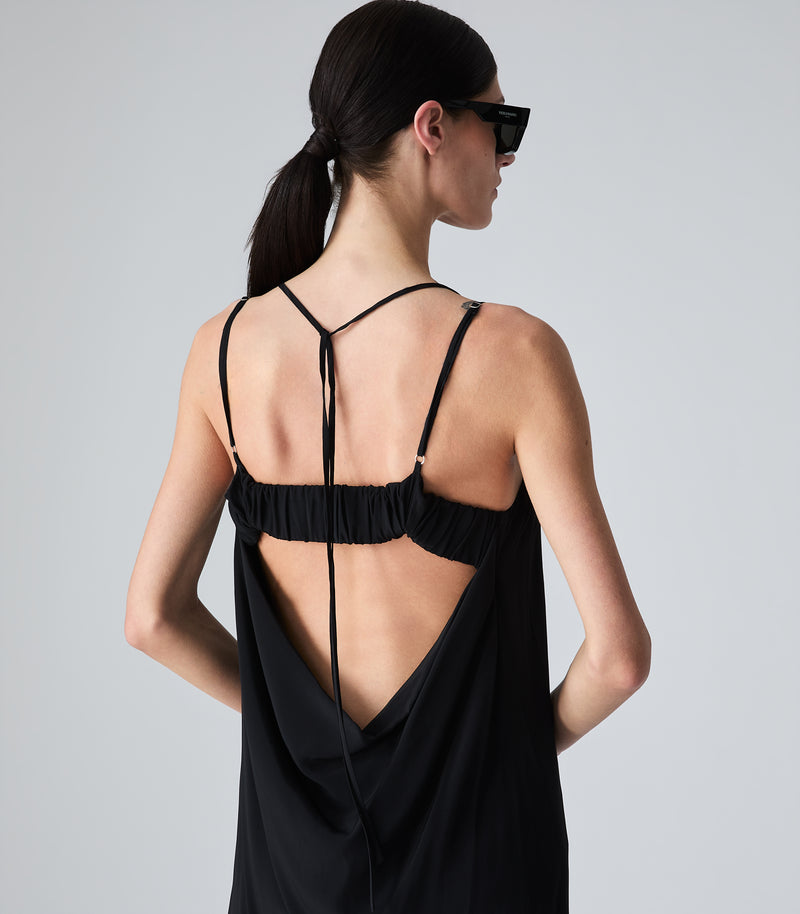 Close-up rear view of a model showcasing the intricate back design of a black slip dress, highlighting a dramatic low open back with fluid cowl draping, a thick ruched horizontal strap, silver hardware loops, and delicate hanging tie strings. The model is turned in profile wearing black sunglasses against a light grey background.