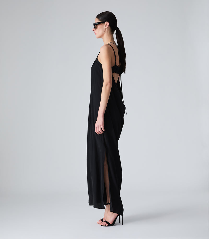Full-length side view of a model showcasing a fluid black maxi slip dress, highlighting a high side leg slit, a loose draped silhouette, and a glimpse of the open-back strap details. The look is styled with black shield sunglasses and black strappy stiletto sandals against a light grey background.