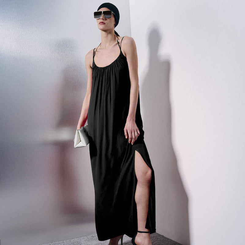 A woman models a black maxi dress with thin spaghetti straps, a gathered smocked bodice, and a high side slit. She is wearing a black beanie, black sunglasses, and holding a small white clutch against a minimalist white and gray studio background.