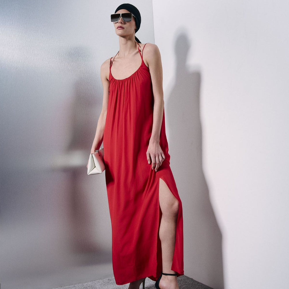 A woman models a red maxi dress with thin spaghetti straps, a gathered smocked bodice, and a high side slit. She is wearing a black head piece, black sunglasses, and holding a small white clutch against a minimalist white and gray studio background.