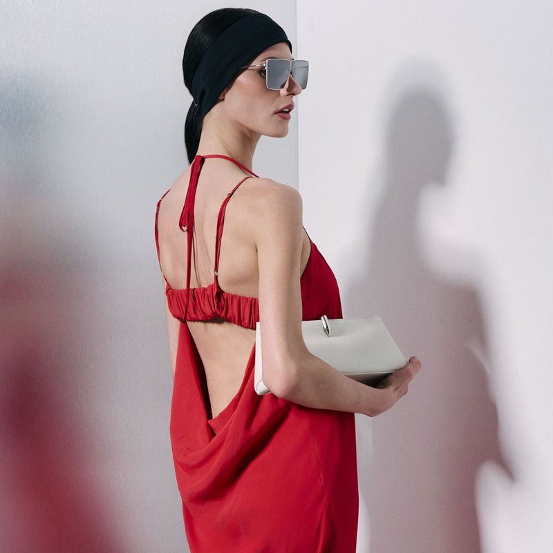 A woman models a red maxi dress with thin spaghetti straps, a gathered smocked bodice, and a high side slit. She is wearing a black head piece, black sunglasses, and holding a small white clutch against a minimalist white and gray studio background.