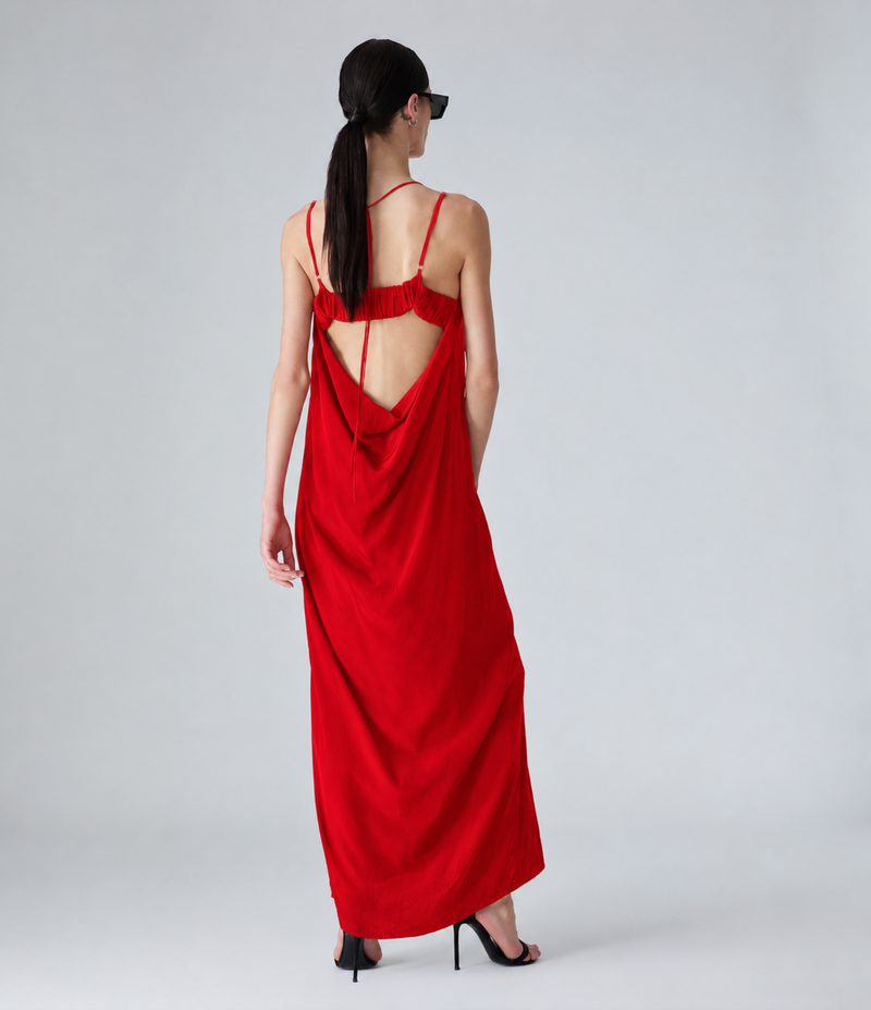 A woman wearing a long red maxi dress with thin straps and a draped open back, photographed from behind against a light gray studio background.