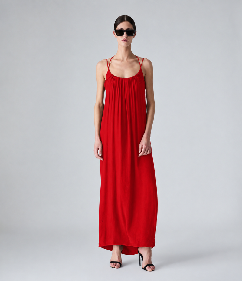 A model wearing a long, bright red spaghetti-strap maxi dress with a gathered neckline, styled with black sunglasses and black high-heeled sandals against a light gray studio background.