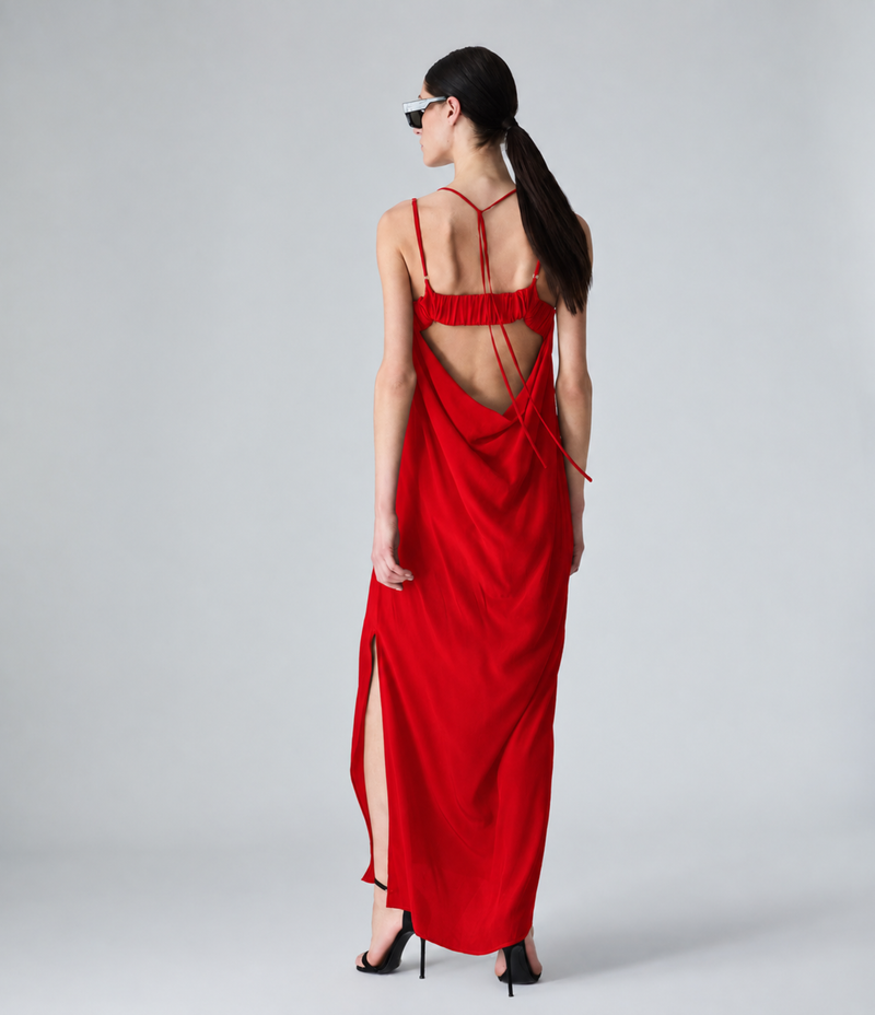 A model wearing a long, bright red spaghetti-strap maxi dress with a gathered neckline, styled with black sunglasses and black high-heeled sandals against a light gray studio background.