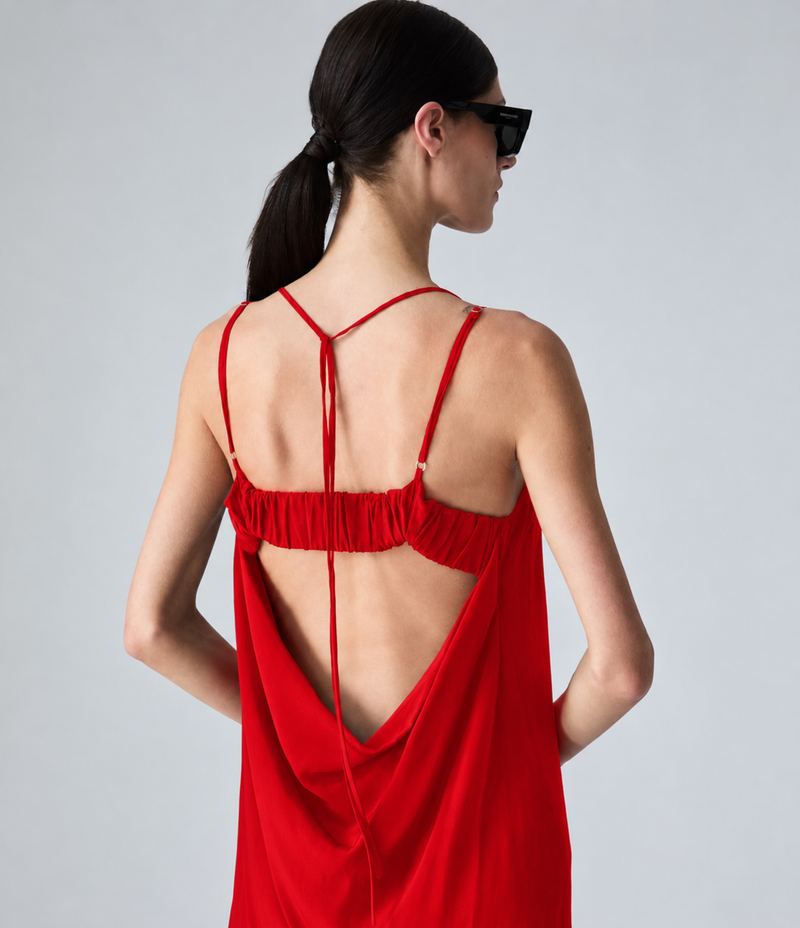 A woman wearing a long red maxi dress with thin straps and a draped open back, photographed from behind against a light gray studio background.
