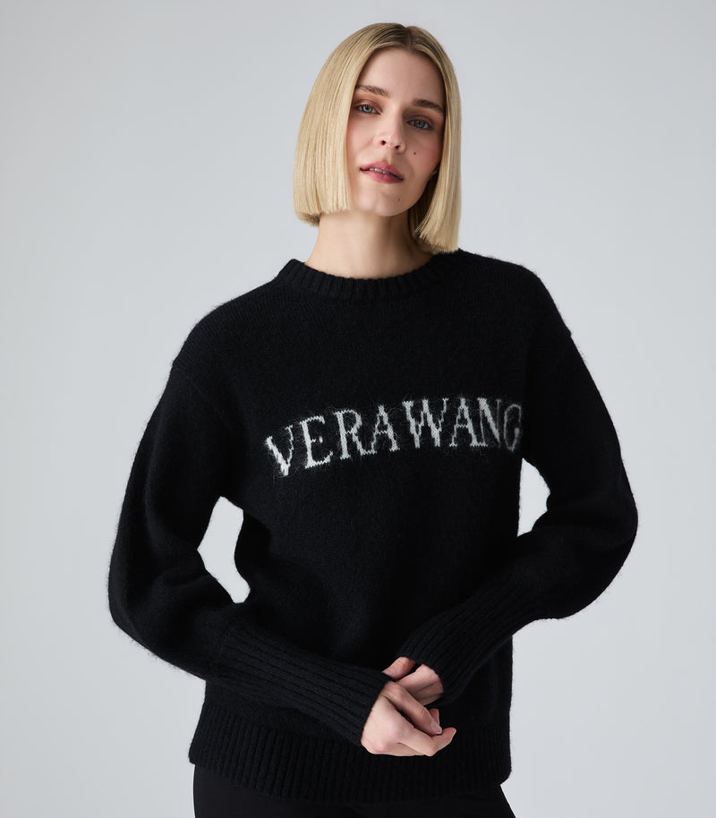 A blonde woman wearing a black knit sweater with 'VERAWANG' embroidered in white across the chest, photographed against a light gray studio background.