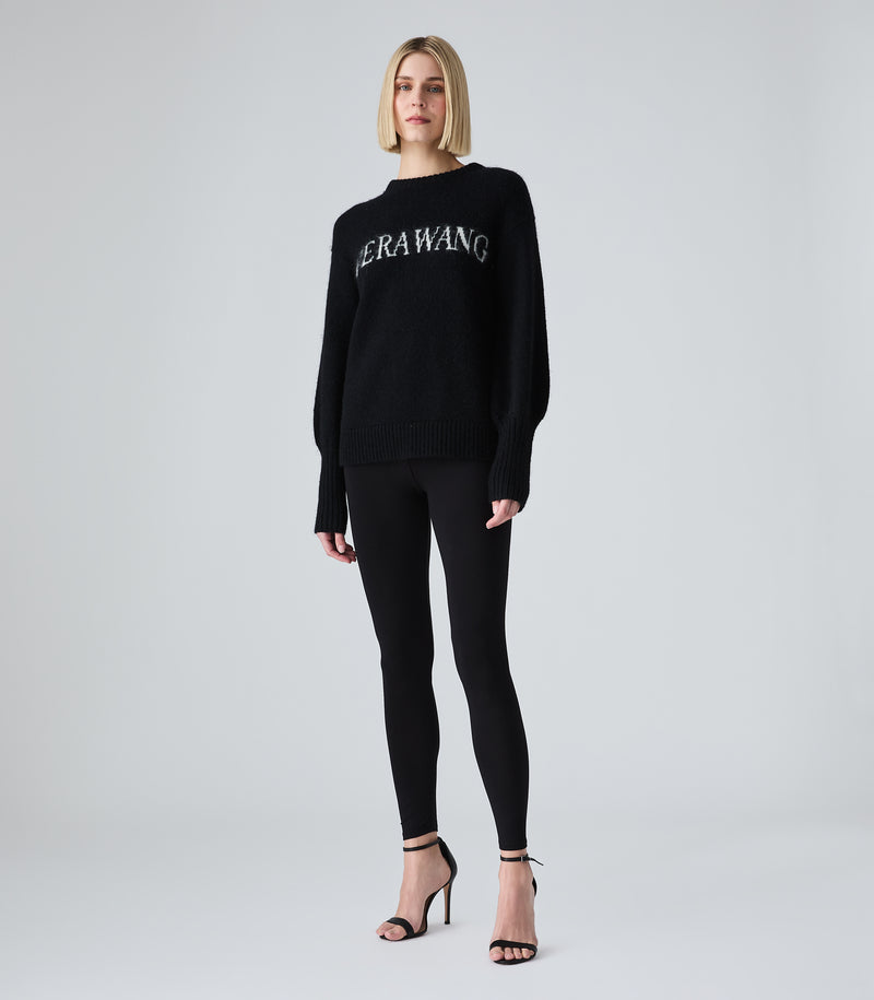 A woman modeling a black knit sweater with 'HERAWANG' embroidered in white across the chest, paired with black leggings and black strappy high heels, against a light gray studio background.