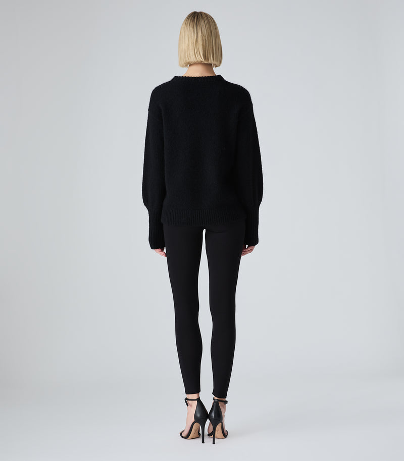 A model viewed from the back wearing a black knit sweater, black leggings, and black high-heeled sandals against a light gray studio background.