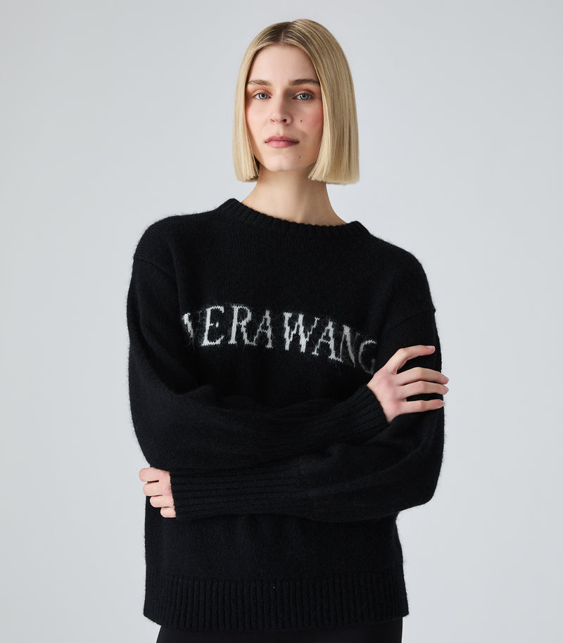 A blonde woman wearing a black knit sweater with 'VERAWANG' embroidered in white across the chest, posed against a light gray studio background.