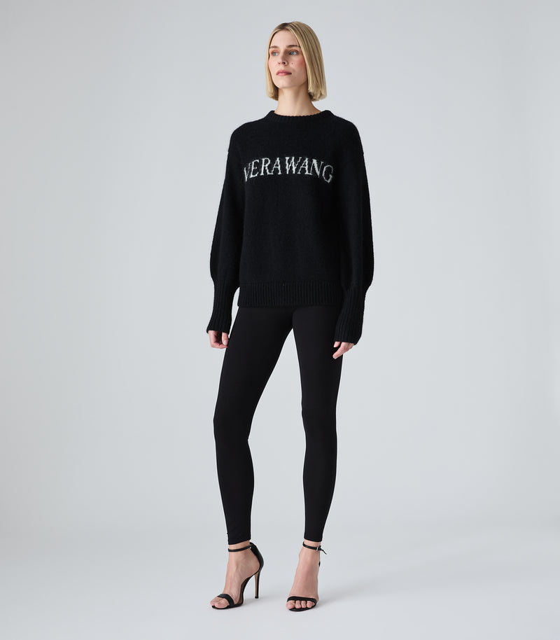 A woman modeling a black knit sweater with 'NERAWANG' embroidered in white across the chest, paired with black leggings and black high-heeled sandals, against a light gray studio background.