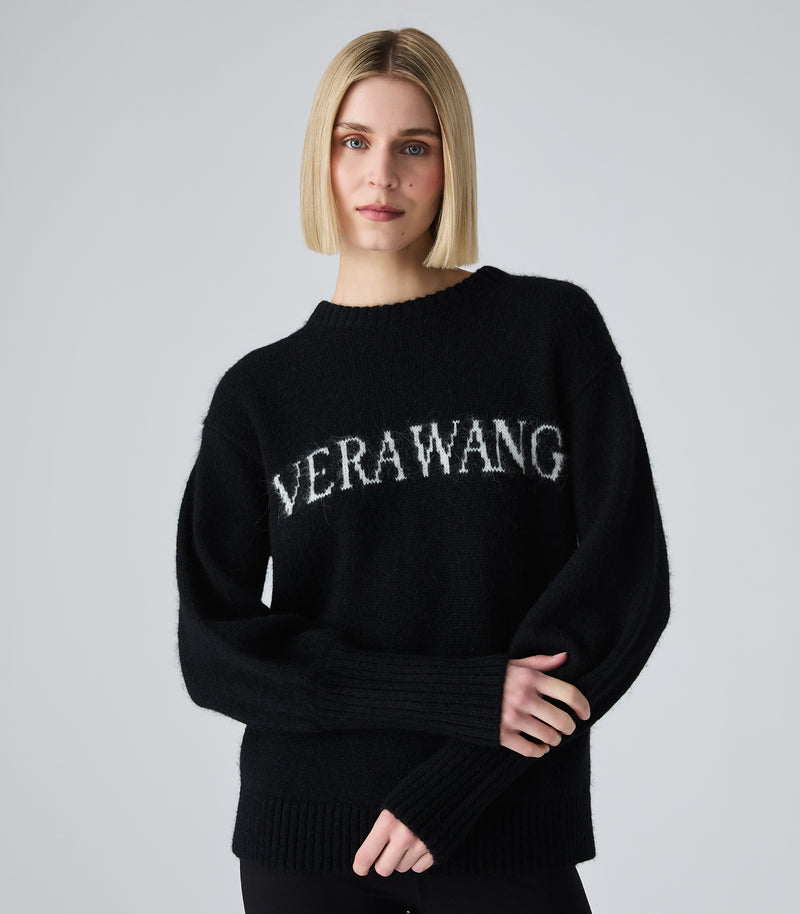 A blonde model wearing a black knit sweater with 'VERAWANG' written across the chest in white textured lettering, photographed against a light gray studio background.