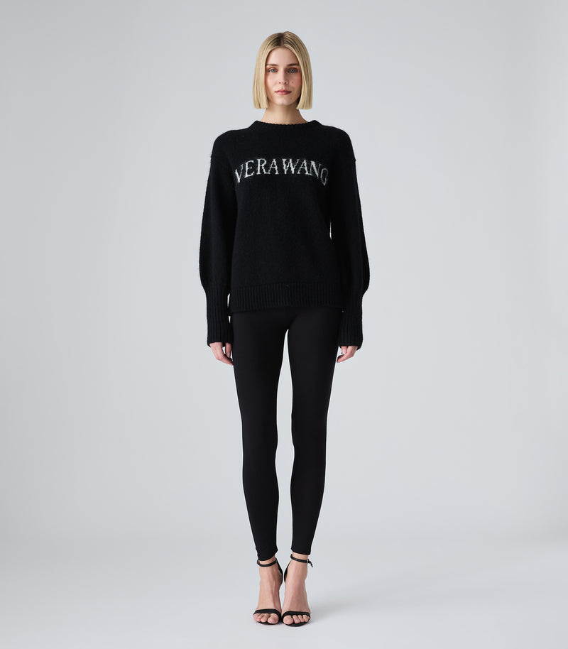 A model stands against a light gray studio background wearing a black knit sweater with 'VERAWANG' embroidered in white across the chest, paired with black leggings and black strappy heels.