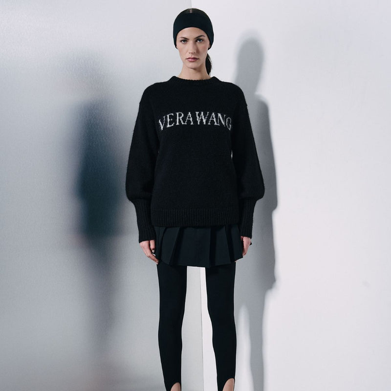 A model stands against a light gray wall wearing a black knit sweater with 'VERAWANG' embroidered in white across the chest, a black beanie, a black pleated mini skirt, and black leggings.