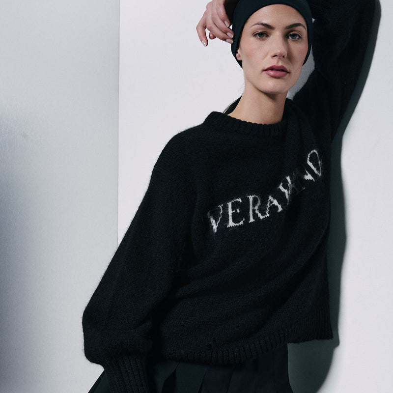 A model wearing a black knit sweater with 'VERA' and 'NIO' in white textured lettering, paired with a black beanie, leaning against a white wall.