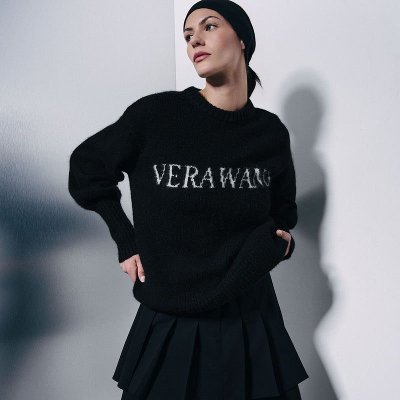 A model wearing a black knit sweater with 'VERAWANG' embroidered in white across the chest, paired with a black pleated skirt and black beanie, set against a gray wall with a soft shadow.