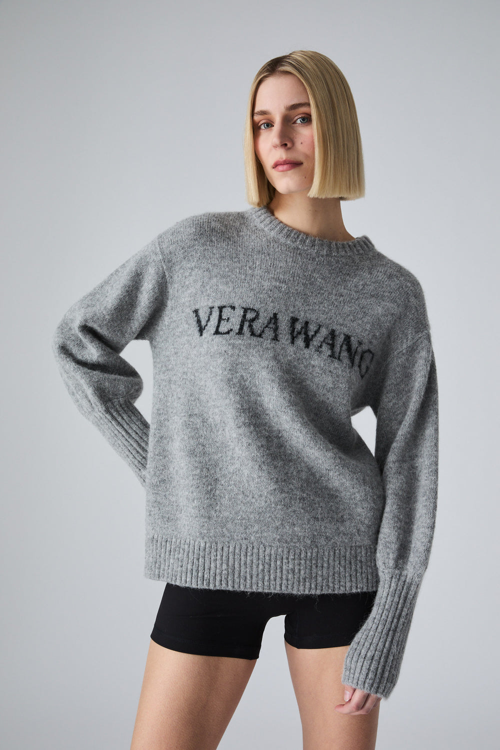 A woman wearing a heather gray knit sweater with 'VERAWANG' embroidered in black across the chest, paired with black shorts, against a light gray studio background.