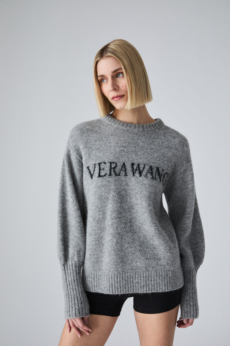 A woman with a blonde bob wearing a heather gray knit sweater with 'VERAWANG' printed across the chest in black, paired with black shorts, against a light gray studio background.